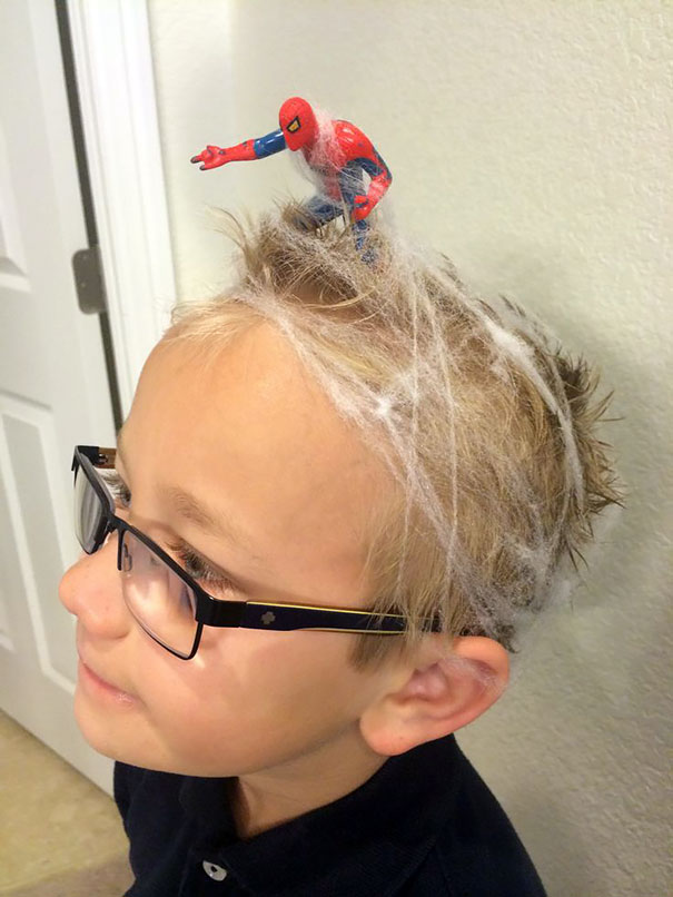 Child with glasses sporting a Crazy Hair Day style, featuring a toy superhero on top with web-like hair decoration.