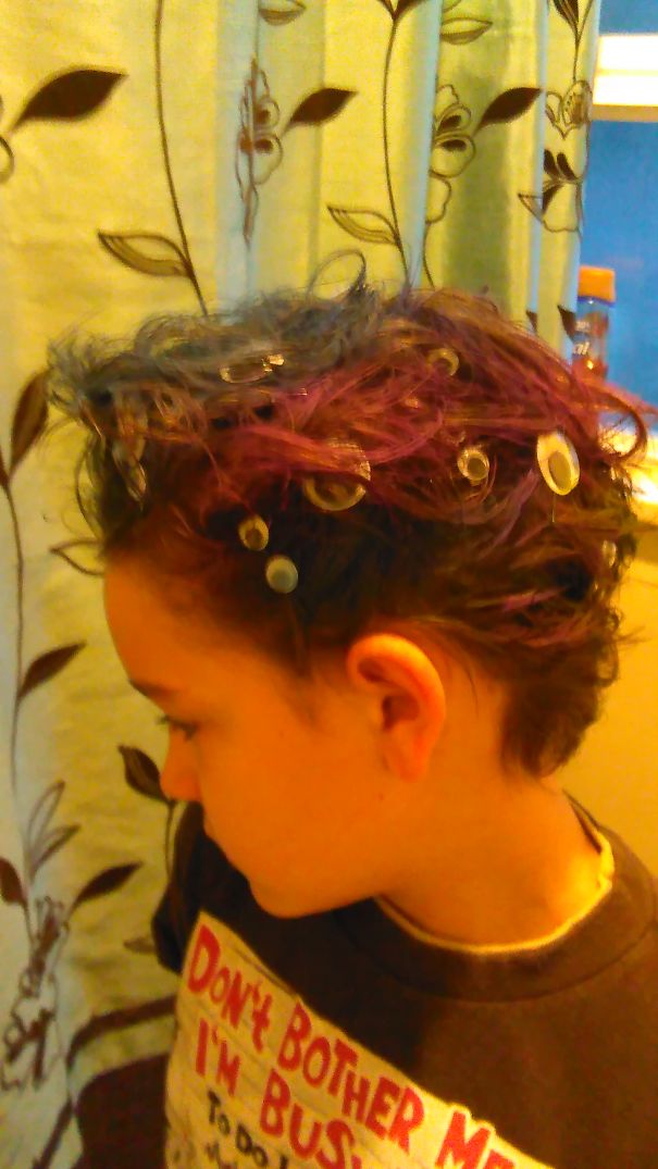 Child with colorful, crazy hair adorned with clear circular clips, in profile view against a floral curtain.