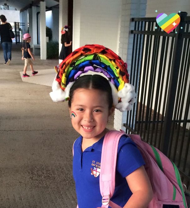 Child with a rainbow-themed crazy hair day style, smiling with a pink backpack and blue shirt.