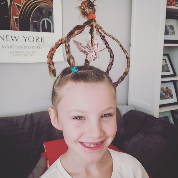 Child with a crown-shaped hairstyle, featuring braided hair and colorful bands, smiling indoors. Crazy Hair Day creativity.