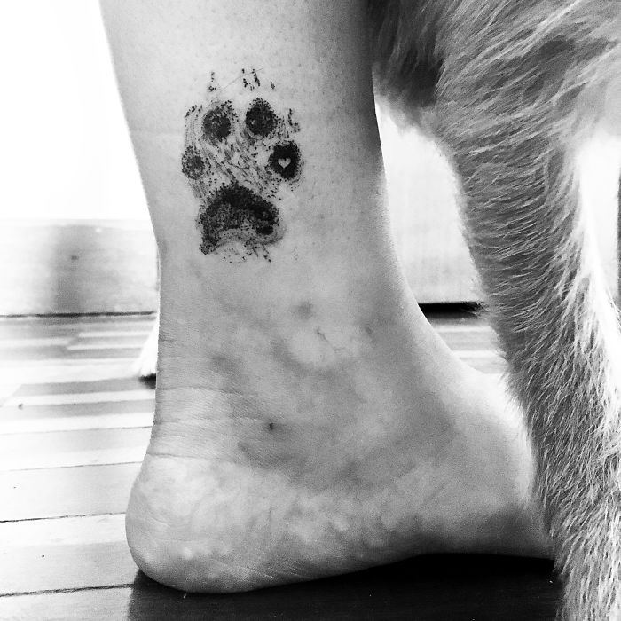 Black paw print tattoo on ankle next to a dog's leg, showcasing unique dog paw prints as tattoo art.