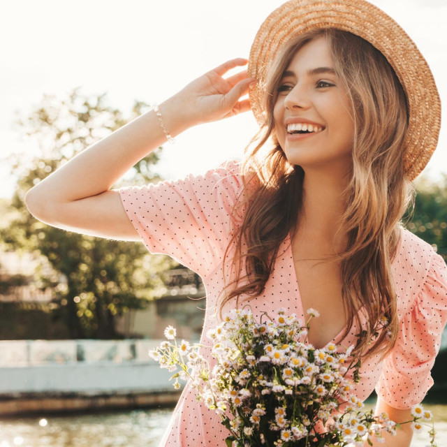 Young beautiful smiling hipster woman in trendy summer sundress. Sexy carefree woman posing on the street background in hat at sunset. Positive model outdoors on embankment. Holding flowers in hands