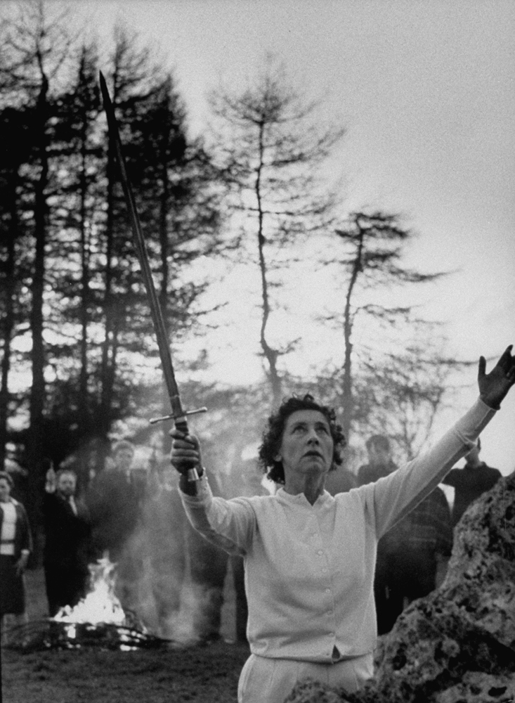 Ray Bone, high priestess of the London witch coven, raises sword and asks 'Mighty Ones of the East' to protect the ritual circle in which they gather near Chipping Norton. Witches behind her hold up knives.