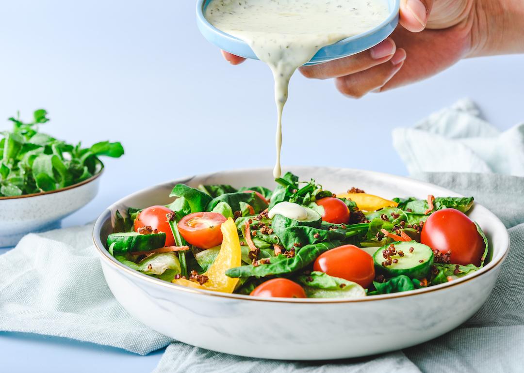 Pouring homemade creamy dressing on a garden salad with cucumbers and tomatoes.