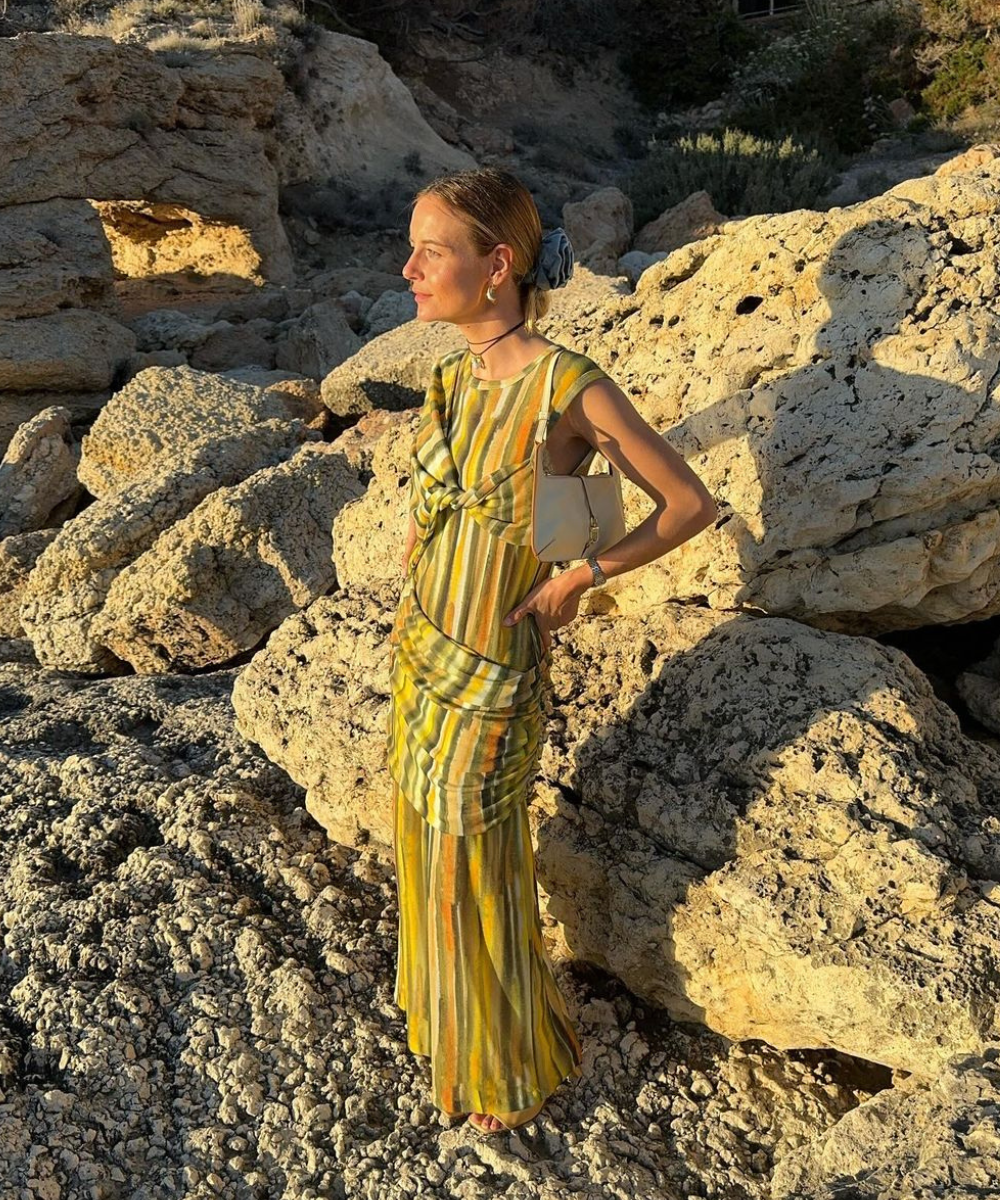 Imagem de uma mulher em um cenário rochoso, vestindo um vestido longo listrado em tons de amarelo e verde. A combinação com pequenos acessórios, incluindo uma bolsa clara e um lenço no cabelo, complementa o visual. O estilo é boho-chic e veranil.