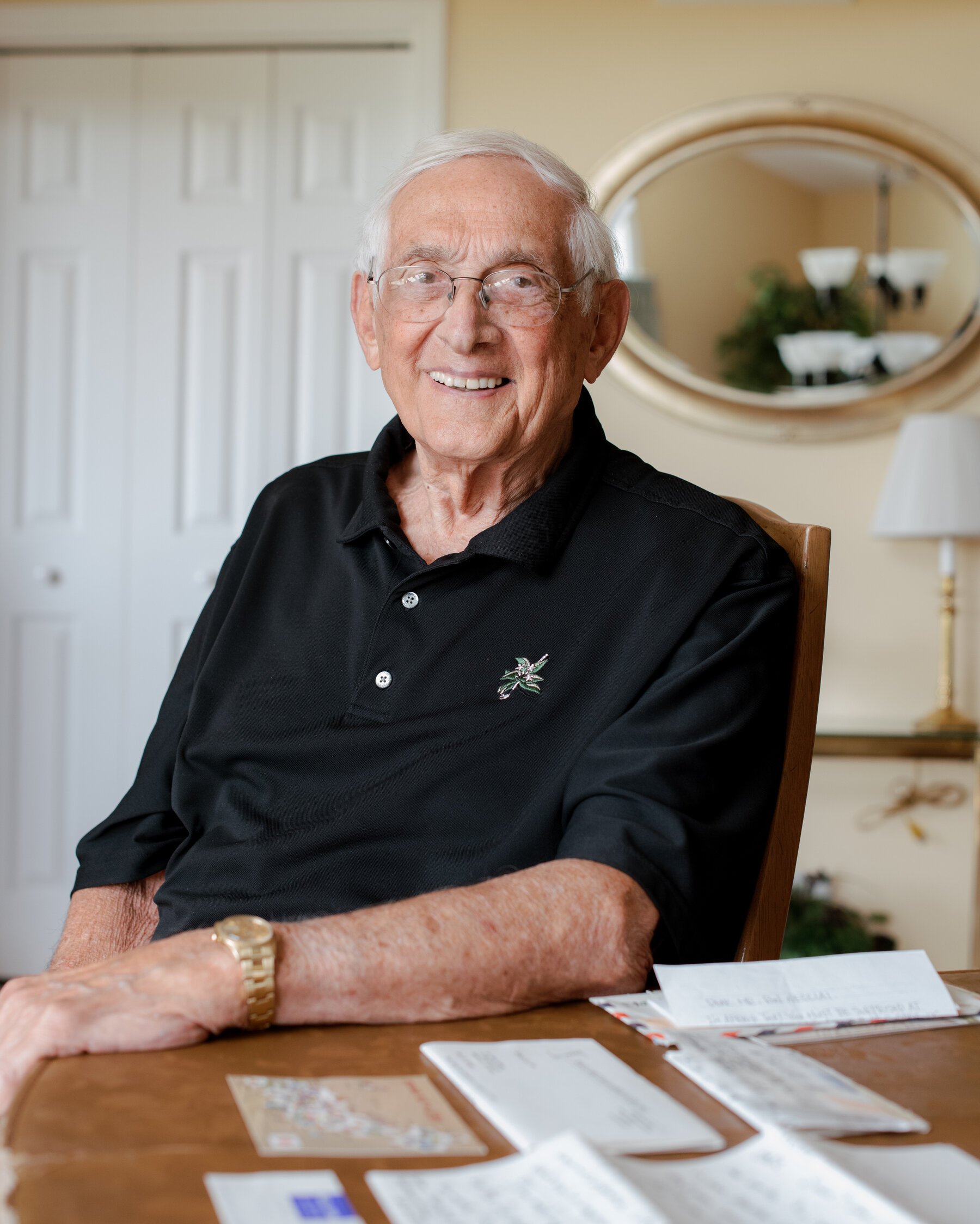 Ron Necciai, wearing a black polo shirt and gold watch, sitting at a table covered with letters in his home.