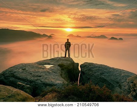 Man stands alone on the peak of rock. Hiker watching to autumn Sun at horizon . Beautiful moment the miracle of nature. Colorful mist in valley. Man hike. Person silhouette stand