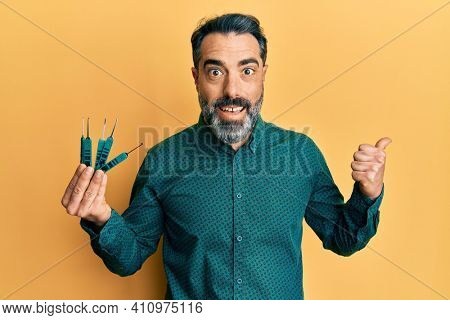 Middle age man with beard and grey hair holding picklock to unlock security door pointing thumb up to the side smiling happy with open mouth 