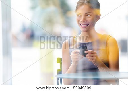 City lifestyle business woman using smartphone on cafe. Young professional female businesswoman on smart phone while sitting indoors in cafe looking out. Multicultural Asian Caucasian girl in her 20s.