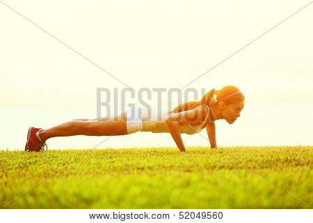 Push ups or press ups exercise by young woman. Girl working out on grass strength training in the glow of the morning sun against a white sky with copyspace. Mixed race Asian Caucasian model.