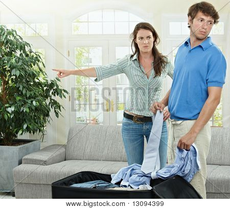 Unhappy couple breaking. Angry woman pointing out, man packing his clothes into suitcase.