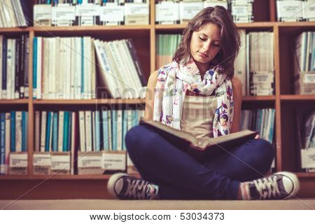 Full length of a female student sitting against bookshelf and reading a book on the library floor