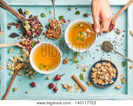 Two cups of healthy herbal tea with mint, cinnamon, dried rose and camomile flowers in spoons and man's hand holding spoon of honey, blue background, top view