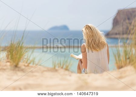 Relaxed woman enjoying sun, freedom and good book an beautiful sandy beach of Balos in Greece. Young lady reading, feeling free and relaxed. Vacations, freedom, happiness, enjoyment and well being.