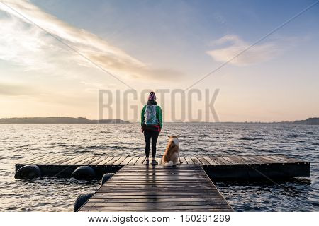 Woman with dog enjoy sunrise and lake relaxing on bridge. Hiker or tourist looking at beautiful morning view with dog friend inspirational landscape on beach. Peaceful people and serene concept.