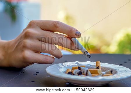 smoking cigarette - woman's hand holding a cigarette over the ashtray full of burnt cigarettes close up view - concept of unhealthy nicotine addiction