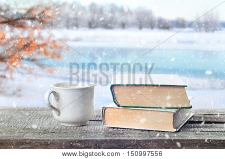 Cup hot coffee or tea cocoa chocolate and book outdoors on wooden table or bench in snowy weather on winter season background. Return to winter time. Pile of books and cup in nature.