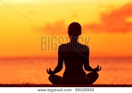 Zen yoga woman doing meditation on beach - wellness concept. Female silhouette relaxing sitting at sunset background meditating at ocean retreat. Summer orange sunrise sky.