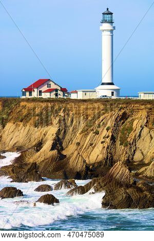 Waves Crashing Onto A Rugged Coastal Bluff With The Historical Point Arena Lighthouse On A Hillside 