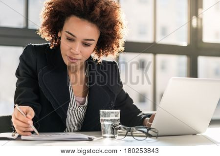 Portrait of African American female expert analyzing printed business report while sitting at desk in the office