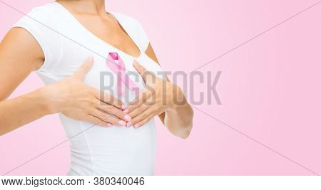 health, medicine and people concept - close up of young woman checking breast with cancer awareness ribbon on chest over pink background