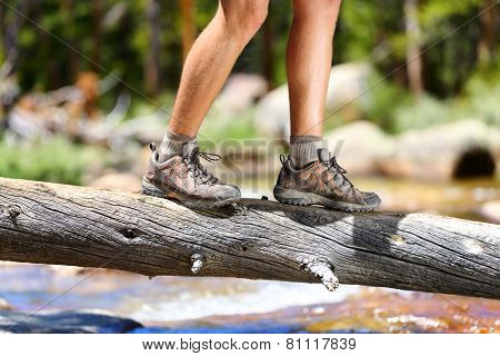 Hiking man crossing river in walking in balance on fallen tree trunk in nature landscape. Closeup of male hiker trekking shoes outdoors in forest balancing on tree. Balance challenge concept.