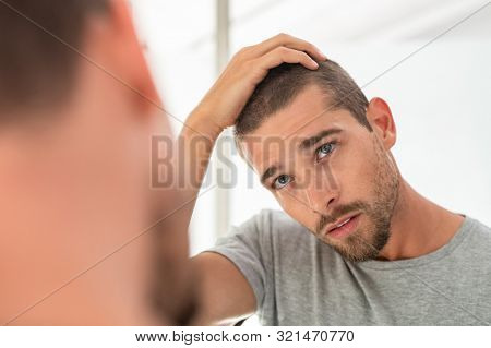 Young unshaven man looking at mirror in bathroom at home. Handsome guy looking at his face in mirror, checking hair and hairline. Man in pijamas concerned with hair loss.