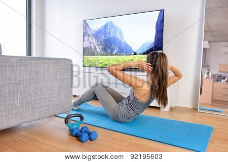 Home workout - woman exercising in front of a flat screen watching a fitness program or exercising during a TV show lying on a yoga mat in front of the sofa in the living room of a house or apartment.