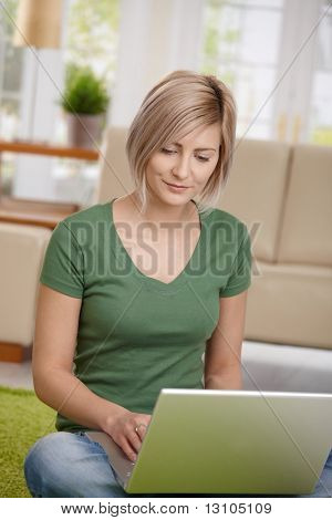 Blond woman sitting on floor at home in living room using laptop computer for browsing internet.