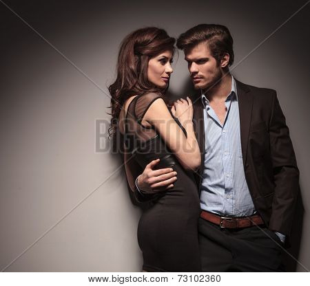 Elegant couple embracing and looking at each other. The man is holding one hand in his pocketOn dark grey background.