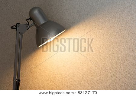 Silver desk lamp or arm lamp in front of a white wall with incandescent type light bulb illuminating the wall.  