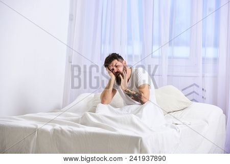 Guy On Sleepy Tired Face Yawning. Man In Shirt Sits On Bed, White Curtains On Background. Nap And Si