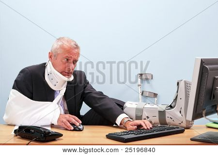 Photo of a mature businessman with multiple injuries sitting at his desk struggling to work on his computer. Good image for health and safety, accident at work or healthcare insurance related themes.