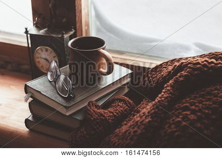 Warm and comfy winter concept. Book, cup of tea and sweater on wooden window sill in old house. Reading and relaxing in cold snowy weather at home. Quiet silent homely scene.