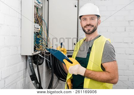 Smiling Handsome Electrician Holding Clipboard Near Electrical Box In Corridor And Looking At Camera