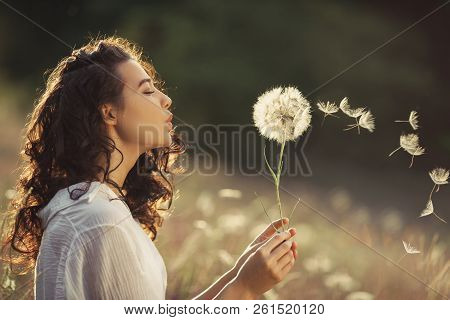 Beautiful Young Woman Sitting On The Field In Green Grass And Blowing Dandelion. Outdoors. Enjoy Nat