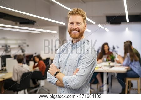 Happy successful businessman standing in office. Portrait of mid adult man entrepreneur standing in open space office with staff working in background. Confident business man looking at camera.