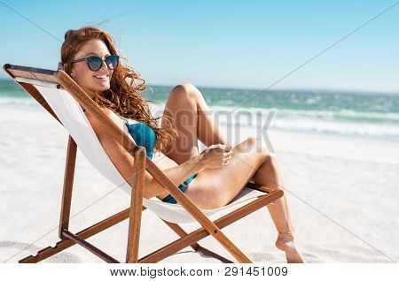 Happy young woman relaxing on deck chair at beach while looking at camera. Mature woman with red hair wearing sunglasses and blue bikini enjoying vacation at beach. Sunbathing and relaxing at sea.