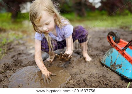 Funny Little Girl Playing In A Large Wet Mud Puddle On Sunny Summer Day. Child Getting Dirty While D