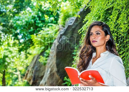 Young Beautiful East Indian American Woman With Long Hair Reading Red Book Outdoor At Central Park, 