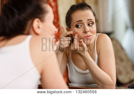 Unhappy young woman finding an acne on a cheek, sits before a mirror.