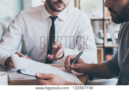 Signing contract. Close-up of confident young man signing some document while another man in shirt and tie sitting close to him and pointing document