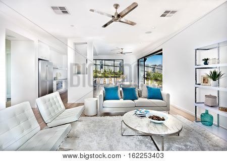 Total white living room decoration interior with the kitchen of a luxury house expressing the color of white within same color walls and furniture. There are round tables and sofas with pillows on the wool carpet next to the shelf.