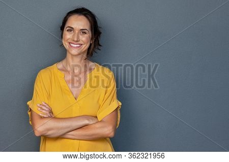 Confident mature woman with crossed arms in casual clothing with copy space. Successful smiling woman with big grin looking at camera. Beautiful positive businesswoman standing against grey background