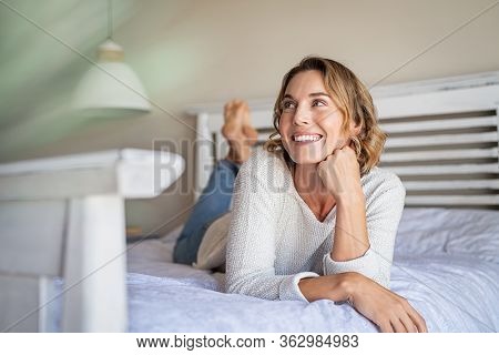 Happy pensive young woman lying and thinking on bed. Thoughtful beautiful girl lying on bed while looking up. Smiling blonde woman contemplating the future at home.