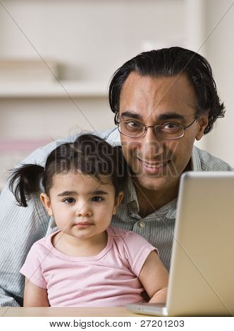 Un padre indio tiene a su hija.  Él sonriendo directamente a la cámara. Hay un ordenador portátil 