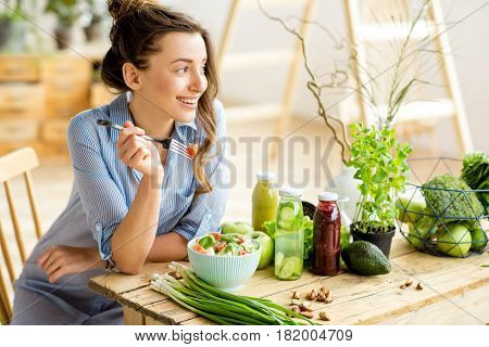 Young and happy woman eating healthy salad sitting on the table with green fresh ingredients indoors