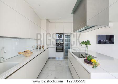 Stylish white kitchen with light tiled floor. There are lockers, tabletop with a sink, kitchen island with a stove and vegetables and kitchen hood, leaves in a vase, fridge with glass door, oven, TV.