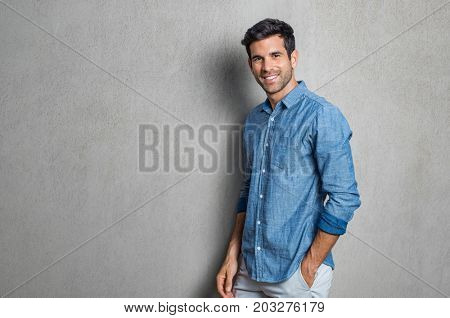 Happy smiling man leaning against grey wall. Portrait of proud mid man isolated on grey background. Young casual hispanic man against grey wall looking at camera.