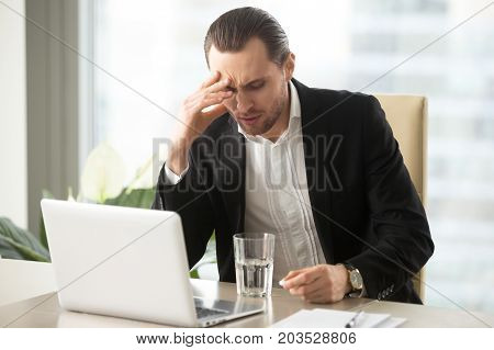 Stressed businessman having severe headache, holding painkiller remedy pill. Sick CEO with head cold, thinking about taking medicine with glass of water. Being ill at work, stress induced migraine.